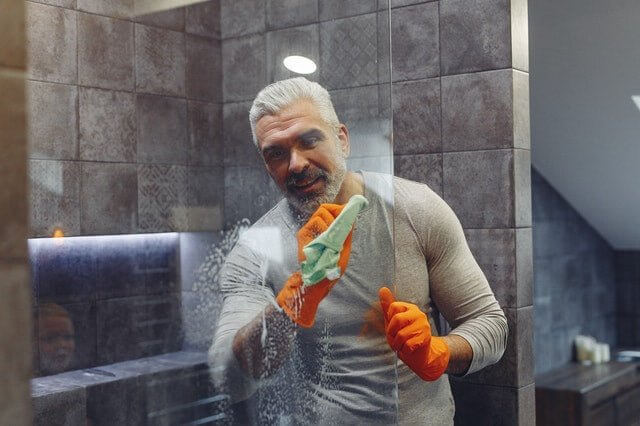 Man wearing orange gloves cleaning a glass shower door with a cloth in a bathroom using quick cleaning tips in 5 minutes