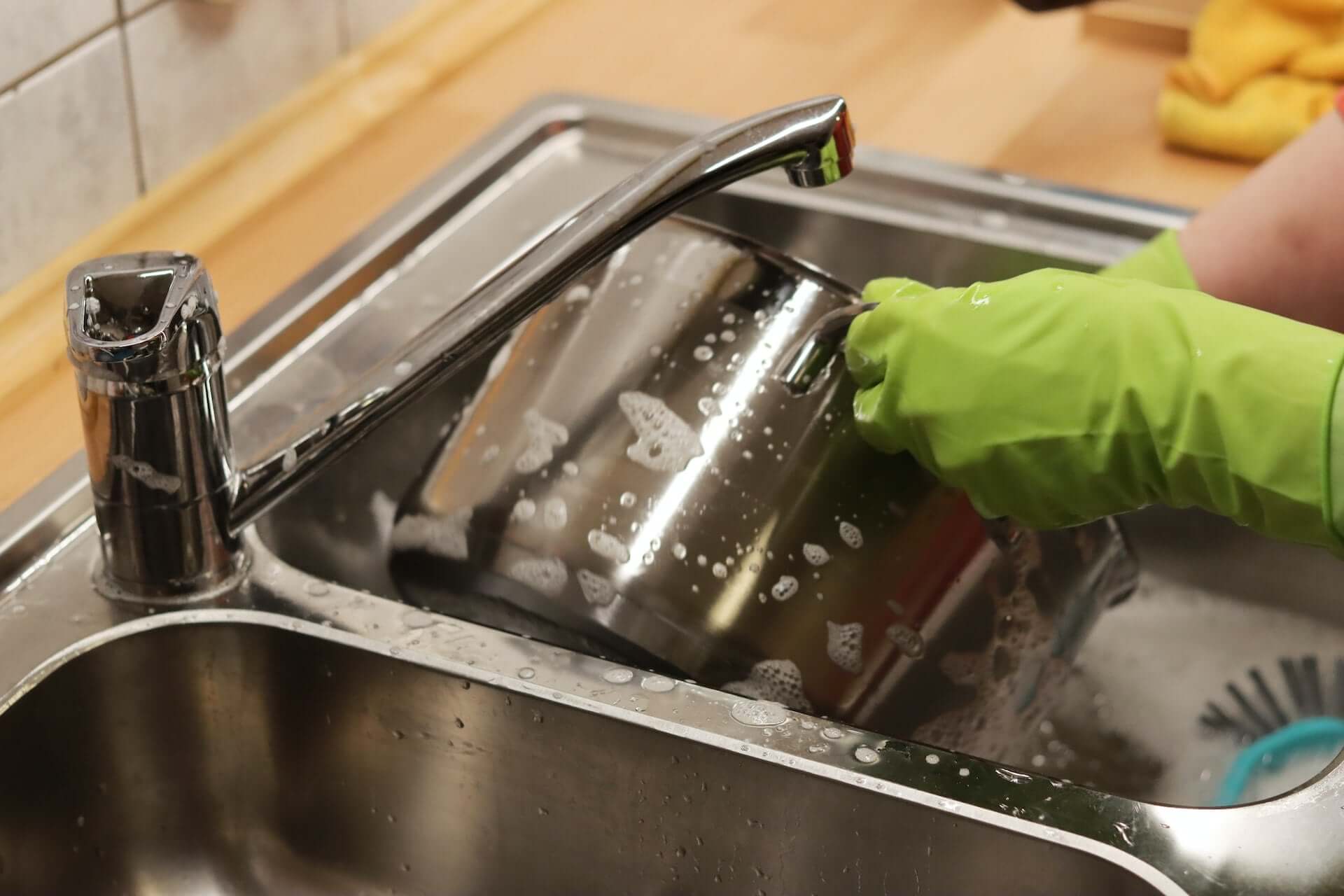 Person wearing green gloves cleaning a stainless steel sink with soapy water and a sponge to clean a stained sink