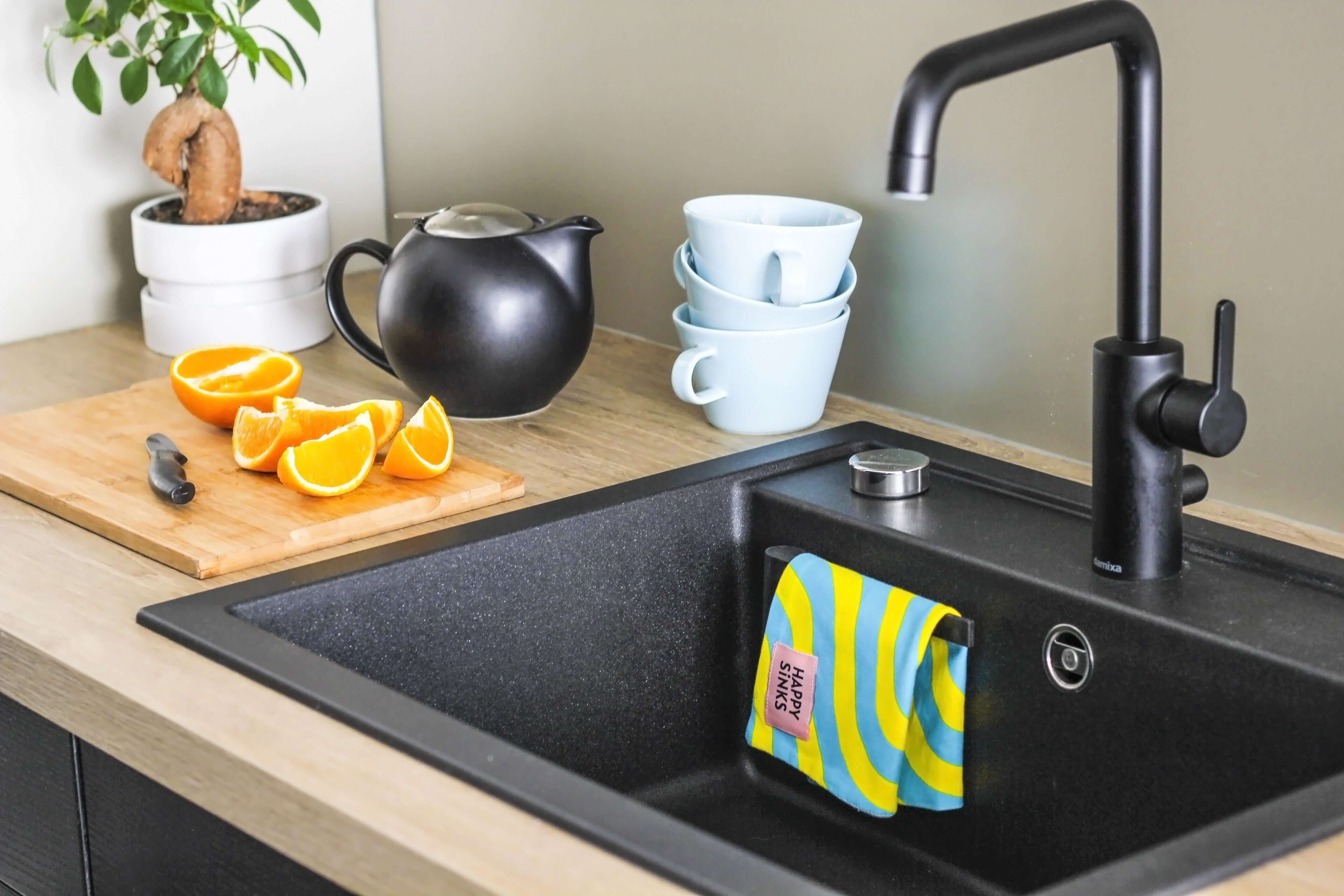 HAPPY SiNKS Cloth Holder attached inside black kitchen sink holding a colorful dish cloth with modern faucet and kitchen countertop with cutting board and teapot.