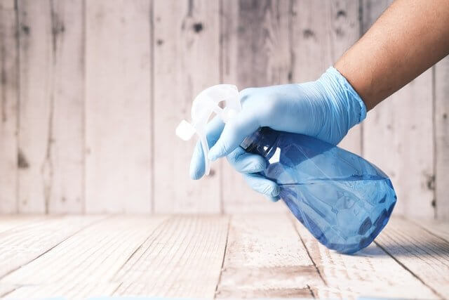Hand wearing blue glove spraying disinfectant from a blue spray bottle on wooden kitchen surface to disinfect the kitchen