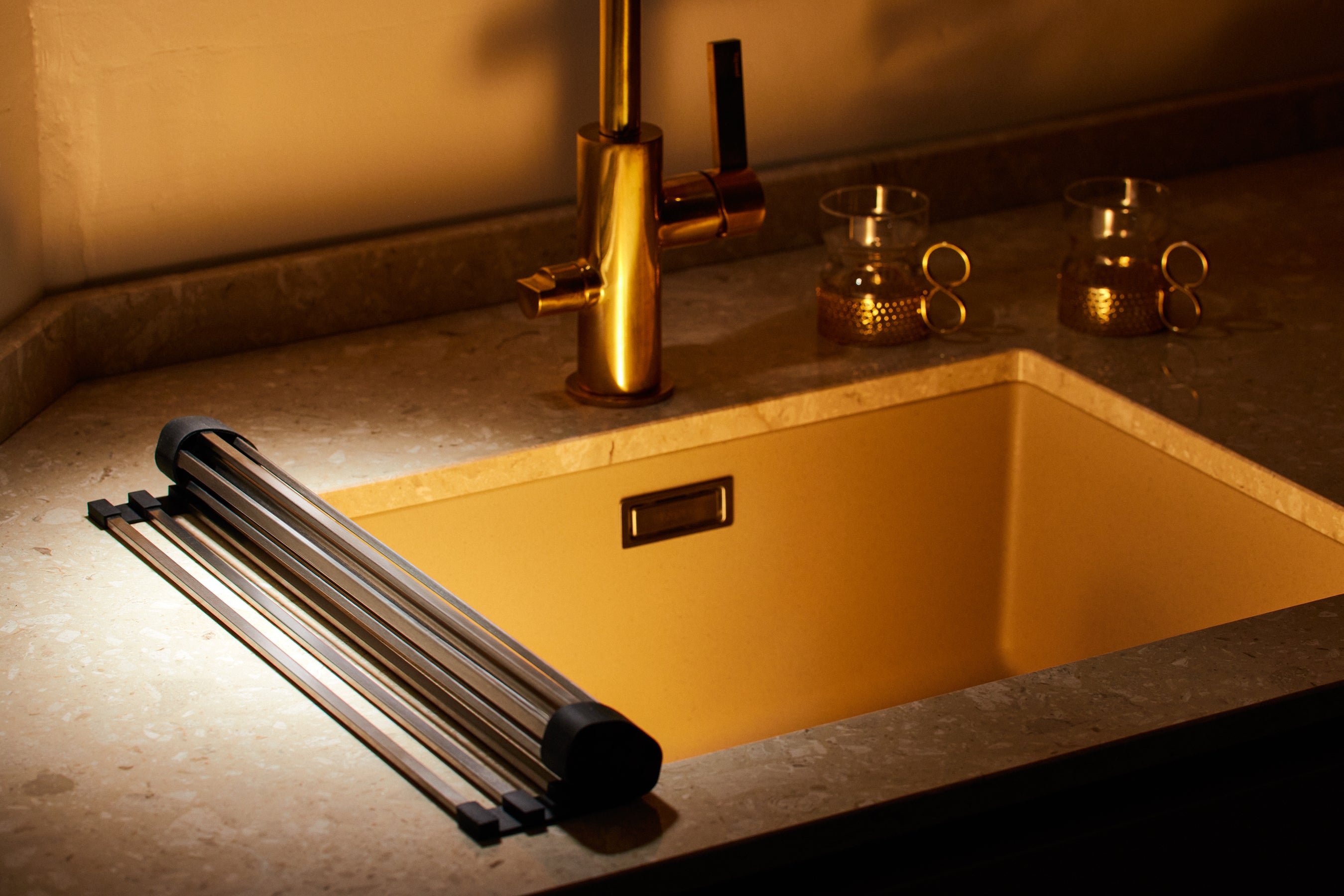 Roll-up stainless steel dish drying rack on a kitchen sink with a faucet and cups nearby.